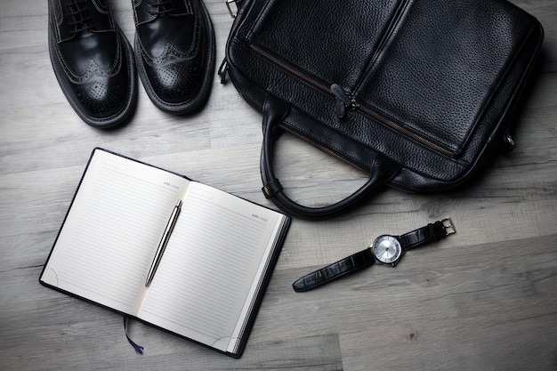 Un sac en cuir noir, un sac en cuir noir, une montre et un sac noir sont posés sur un plancher en bois.