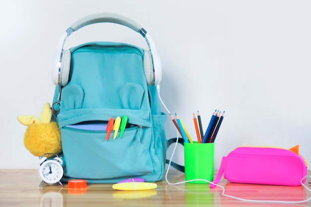 Un sac à dos d'école amusant avec des écouteurs, une trousse lumineuse, un porte-clés moelleux et un verre avec des crayons de couleur et un réveil sur une table en bois. concept de retour à l'école.