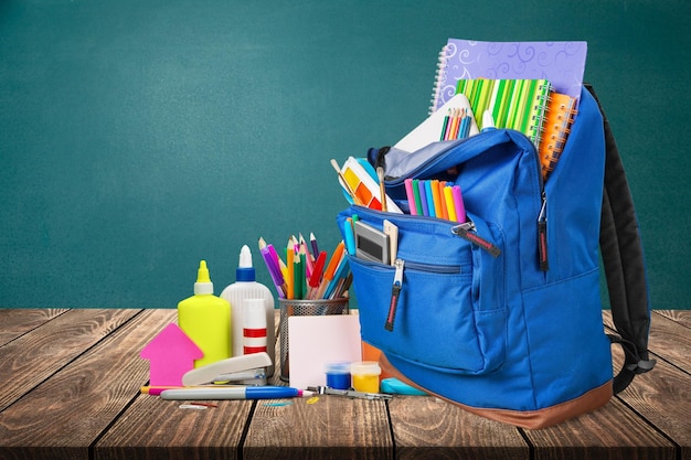 Sac à dos élégant avec différentes fournitures scolaires sur une table en bois blanche au tableau vert.