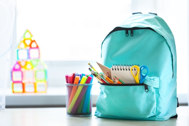 Sac à dos scolaire bleu avec des fournitures scolaires sur la table dans la chambre des enfants en face de la fenêtre en journée ensoleillée