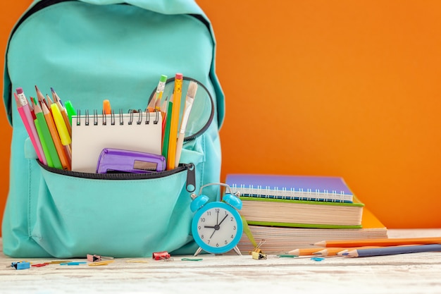 Sac à dos scolaire avec différentes fournitures et réveil sur fond orange.