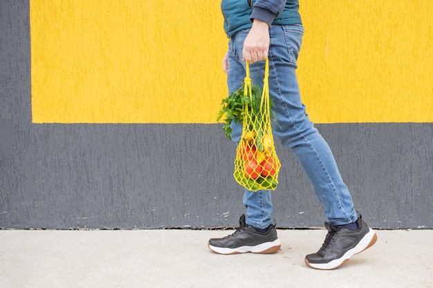 Sac à ficelle jaune avec concombres, tomates, bananes et herbes dans les mains de l'homme en jeans. Photo lumineuse dans les tons rouges, jaunes et verts. Développement durable, zéro déchet, concept sans plastique.