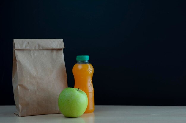 sac en papier avec la collation pour l'école avec une pomme verte et une bouteille de jus d'orange sur le bureau sur un fond noir du tableau avec un endroit pour l'inscription