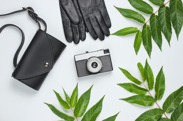 Sac de taille en cuir, gants, appareil photo rétro sur fond blanc avec des feuilles tropicales vertes. Vue de dessus