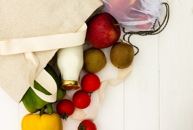 Sacs à provisions en coton avec des légumes et des fruits et une bouteille de lait en verre sur un fond en bois blanc. Vue de dessus. Espace de copie. Concept zéro déchet et respectueux de l'environnement.