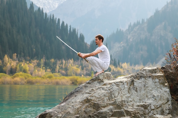 Un sage est assis sur une pierre et tient une épée dans ses mains. Fond de lac de montagne.