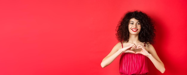 Saint Valentin fille romantique avec une coiffure bouclée en robe de soirée souriante et montrant le signe du cœur sa