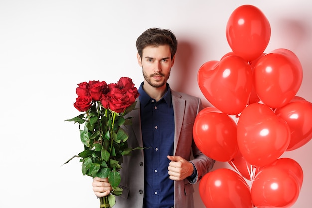 La Saint-valentin. Homme Beau Et Confiant, Rendez-vous En Costume, Tenant Un Bouquet De Roses Rouges Et Des Ballons Coeur, Debout Avec Des Cadeaux Pour Amoureux Sur Fond Blanc.