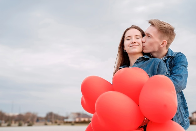 La Saint-Valentin. Jeune couple aimant étreindre et s'embrasser tenant des ballons en forme de coeur rouge à l'extérieur