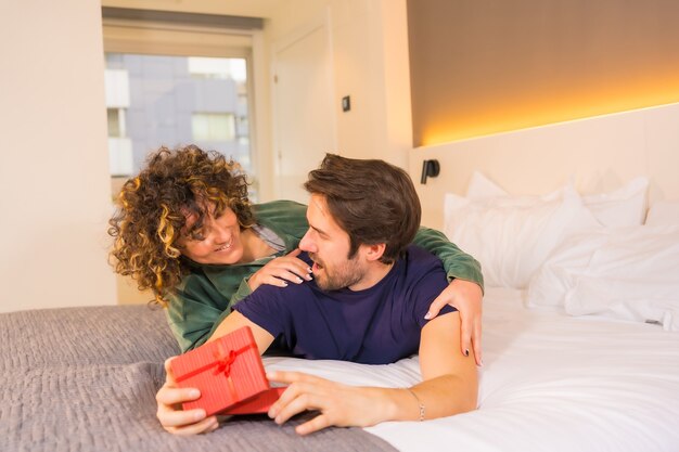 La Saint-Valentin, à un jeune couple caucasien en pyjama sur le lit, la mariée donne une belle surprise au petit ami avec un cadeau. Avec un espace copier-coller à droite