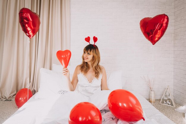 Saint Valentin, Journée de la femme. Jeune fille caucasienne assise dans son lit et célébrant la Saint-Valentin tenant un ballon en forme de coeur dans ses mains