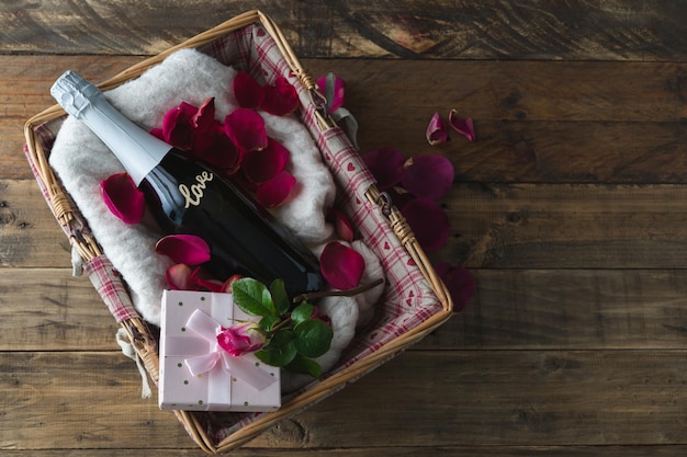 La Saint-Valentin. Panier cadeau avec bouteille de champagne avec inscription LOVE sur la bouteille et coffret cadeau avec rose. Décoration avec pétales de rose et rouge sur fond en bois. Espace de copie.