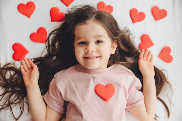 Saint Valentin une petite fille allongée à la maison sur un lit blanc parmi les coeurs souriant et riant de bonheur félicitant pour les vacances