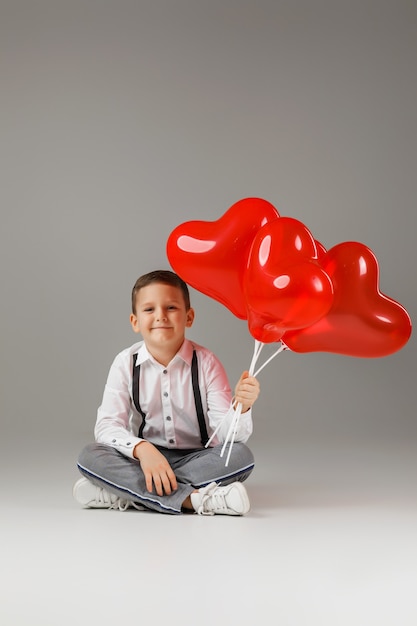 La Saint-Valentin. souriant enfant élégant garçon tenant des ballons en forme de coeur rouge et assis sur le sol
