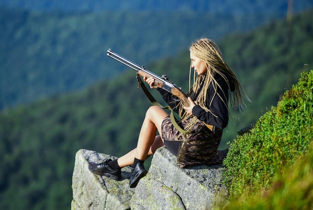 Saison de chasse Fond de paysage de montagnes de chasseur Guerrier sexy Femme séduisante cheveux longs joli visage tenir le fusil pour la chasse Fille amazone s'asseoir sur la falaise préparer le pistolet pour la chasse Concept de visée