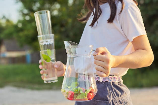 Saison estivale, chaleur, boissons non alcoolisées. Girl wearing hat avec cruche avec boisson froide à la menthe aux fraises maison naturelle et verres vides, fond de style campagnard