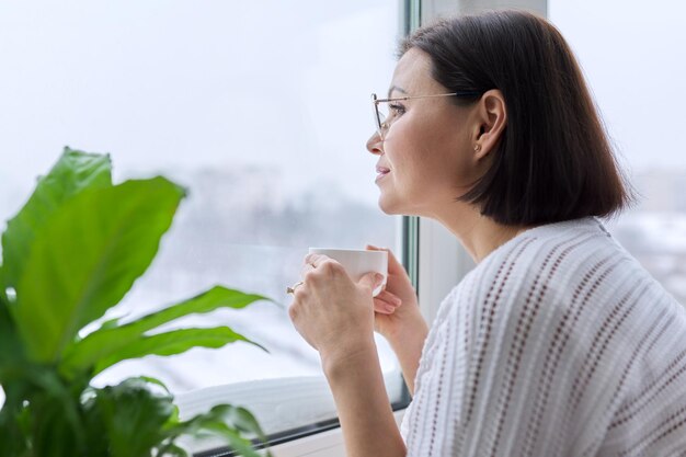 Saison hiver jour enneigé femme souriante d'âge moyen avec une tasse de café regardant par la fenêtre