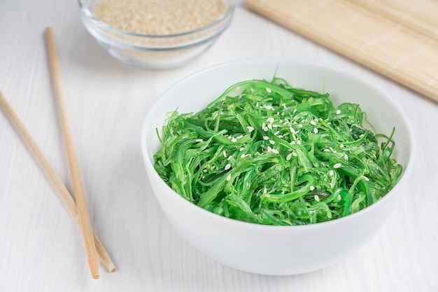 Salade d'algues chuka wakame vert aux graines de sésame dans un bol avec des baguettes sur table en bois blanc