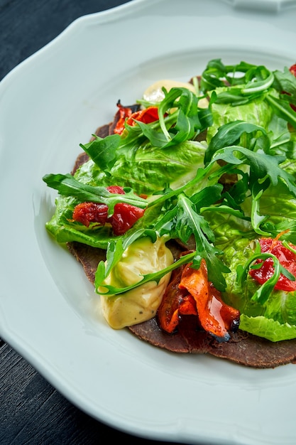 La salade apéritive italienne traditionnelle est le vitello tonnato. Boeuf émincé avec roquette, tomates et pesto, servi dans une assiette blanche sur une surface sombre