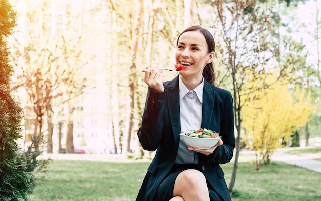 Salade au déjeuner d'affaires Portrait d'une belle femme d'affaires moderne et souriante dans un costume noir avec une assiette de salade de légumes dans les mains qu'elle mange dans la rue près du travail