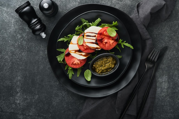 Salade caprese Salade caprese italienne avec tranches de tomates fromage mozzarella roquette basilic huile d'olive dans une assiette noire sur de vieux carreaux de brique fond noir Délicieuse cuisine italienne Vue de dessus