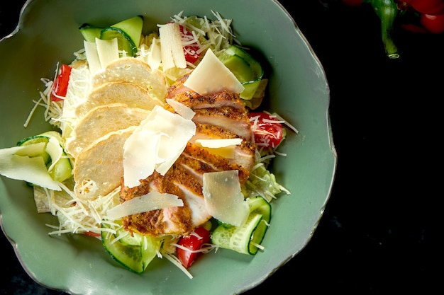 Salade César classique avec croûtons, parmesan, poulet grillé, servie dans un bol vert sur une table en marbre foncé. Nourriture de restaurant.