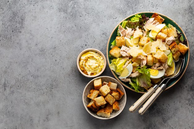 Salade César fraîche avec salade de laitue, poitrine de poulet, œufs durs et croûtons dans un bol en céramique avec vinaigrette sur le côté sur une table grise. Salade saine classique, vue de dessus, espace pour le texte