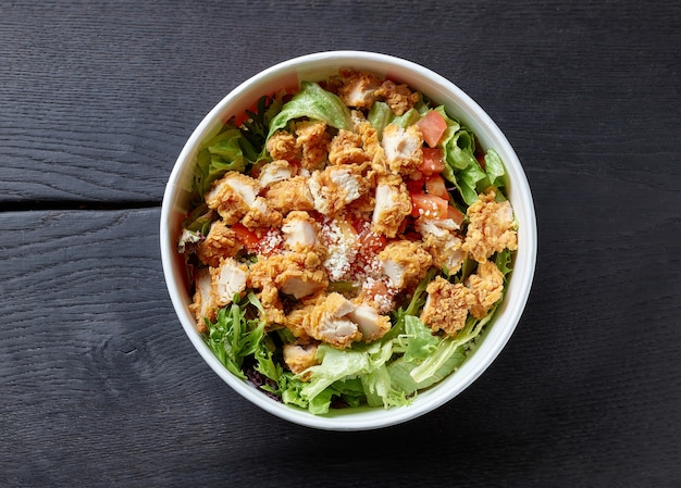 Salade césar fraîche avec de la viande de poulet frit dans un récipient à emporter en papier sur une table en bois noir, vue de dessus