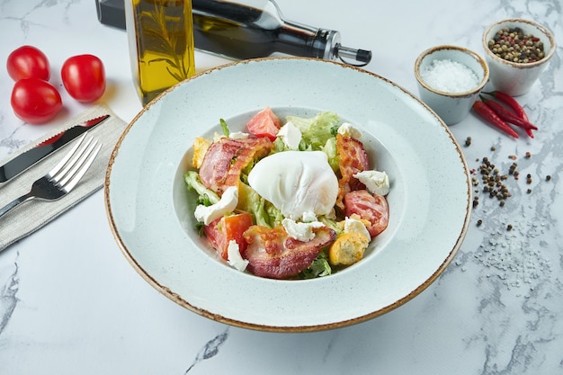 Salade césar ou lardon appétissante avec croûtons, bacon, fromage feta, tomates et œuf poché dans une assiette bleue sur une surface en marbre