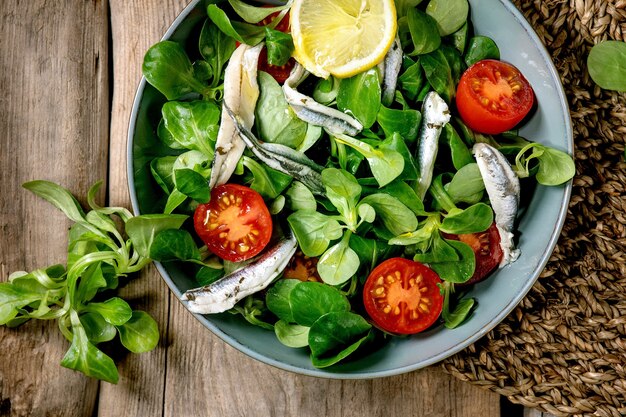 Photo salade de champ vert avec filet d'anchois ou de sardines marinés et tomates cerises, servi dans un bol bleu avec du citron et de l'huile d'olive sur une serviette en paille sur une vieille surface en bois