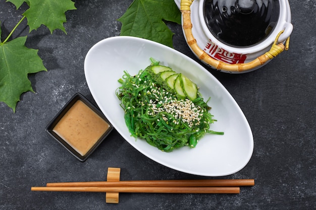 Salade de chuka aux concombres, graines de sésame et sauce, dans une assiette blanche, avec une théière japonaise, des baguettes et des feuilles d'érable, sur du béton noir