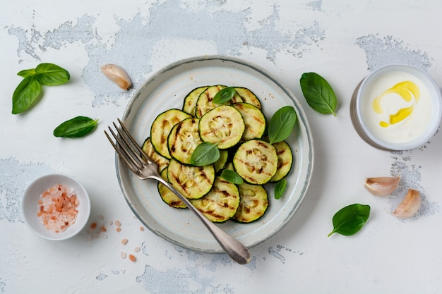 Salade de courgettes grillées avec feuilles de basilic, sauce au yogourt et pain frit dans une simple plaque en céramique sur une surface en béton blanc