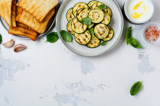 Salade de courgettes grillées avec feuilles de basilic, sauce au yogourt et pain frit dans une simple plaque en céramique sur une surface en béton blanc