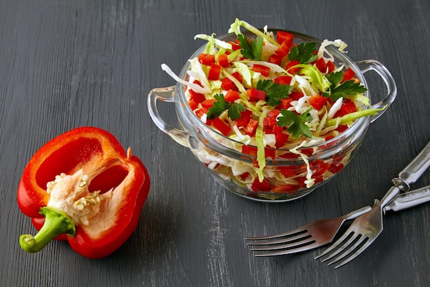 Salade d'été légère de paprika de chou blanc frais et de persil assaisonné d'huile et de citron dans un bol de verre et des couverts sur une table en bois noir