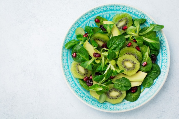 Salade de feuilles de bébé épinards, cresson, kiwi, avocat et grenade dans une plaque en céramique bleue sur une surface en bois clair blanche. Mise au point sélective. Vue de dessus.