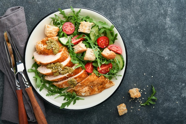 Salade fraîche avec filet de poulet grillé laitue roquette épinards tomates concombre et sauce pesto graines de sésame à l'huile d'olive dans un bol blanc sur fond d'ardoise noire Menu déjeuner sain Vue de dessus