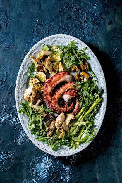 Salade de fruit de mer. Tentacules grillés cuits de poulpe, de sardines et de moules sur une assiette en céramique bleue, servis avec une salade de roquette, des courgettes et des asperges sur une surface de texture bleue. Vue de dessus, mise à plat