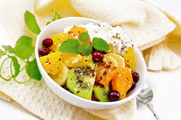 Salade de fruits avec banane, orange, kiwi, canneberges et citrouille au four, crème fouettée, saupoudrée de chocolat et noix de coco à la menthe dans un bol sur une serviette sur fond de planche de bois