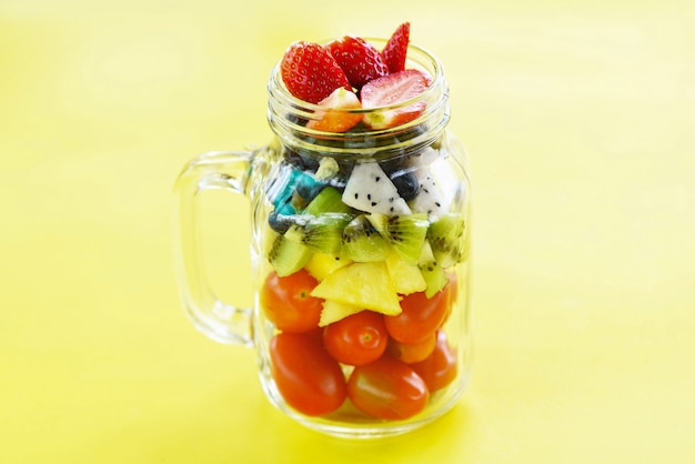 Salade de fruits dans un bocal de verre fruits frais d&#39;été frais et légumes nourriture biologique saine fraises kiwi myrtilles fruit du dragon tropical tomate ananas sur jaune