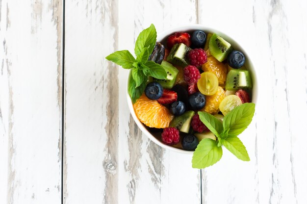 Salade de fruits frais et de baies, feuilles de menthe sur fond de bois blanc. vue de dessus.