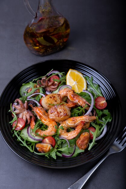 Salade de fruits de mer, roquette, tomates, concombres, oignon rouge et citron sur une assiette noire.