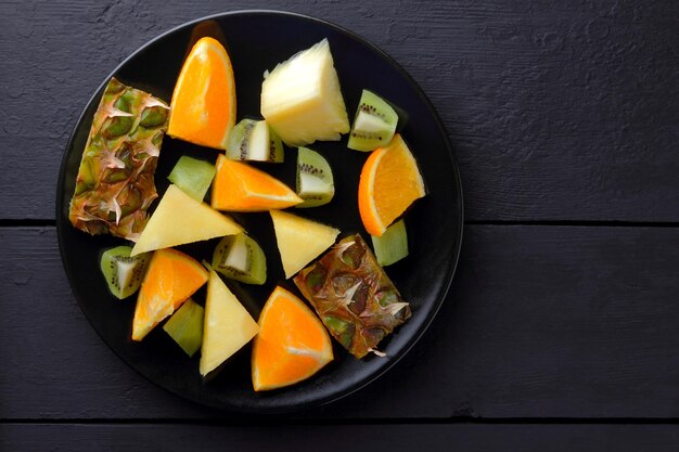 Salade de fruits sur plaque noire Hineapples oranges et kiwi sur fond sombre Salade végétarienne de fruits tropicaux Vue de dessus Espace de copie