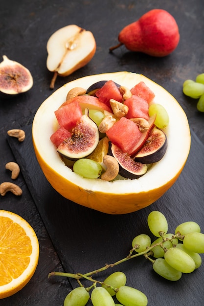 Salade de fruits végétariens de pastèque, raisins, figues, poire, orange, noix de cajou sur ardoise sur fond de béton noir. Vue de côté, gros plan.