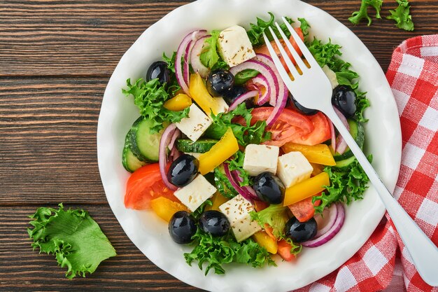 Salade grecque traditionnelle de concombre frais, tomate, poivron, laitue, oignon rouge, fromage feta et olives à l'huile d'olive sur plaque blanche. Alimentation saine, vue de dessus.