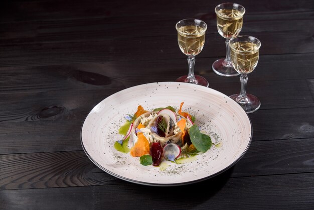 Salade de hareng, mayonnaise, oignon, garnie de chips de citrouille, oignons bleus, fleurs sur une assiette blanche sur une table en bois sombre avec des piles d'alcool.