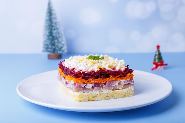 Salade de hareng sous un manteau de fourrure. Salade russe traditionnelle à plusieurs couches de hareng, betteraves, pommes de terre, carottes et œufs. Gros plan, fond bleu, sapin de Noël miniature.