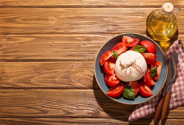 Salade italienne aux tomates fraîches, fromage burrata, basilic servi sur une assiette en céramique avec une bouteille d'huile d'olive sur fond de bois rustique d'en haut, collation saine ou déjeuner avec un espace pour le texte