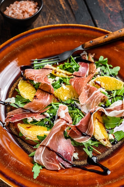 Salade de jambon au jambon, parmesan, roquette et mandarine sur une assiette. Table en bois foncé. Vue de dessus.