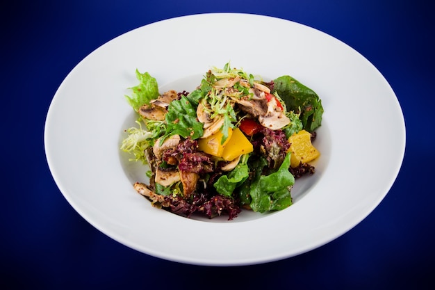 Salade de légumes aux champignons, poivrons, oignons, laitue et sauce aux graines de moutarde. Vue de dessus à plat sur une plaque blanche sur fond bleu.