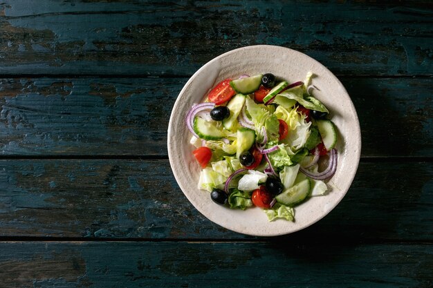 Salade de légumes classique avec tomates, concombre, oignon, feuilles de salade et olives noires dans une assiette en céramique blanche. Fond en bois foncé. Mise à plat, espace de copie